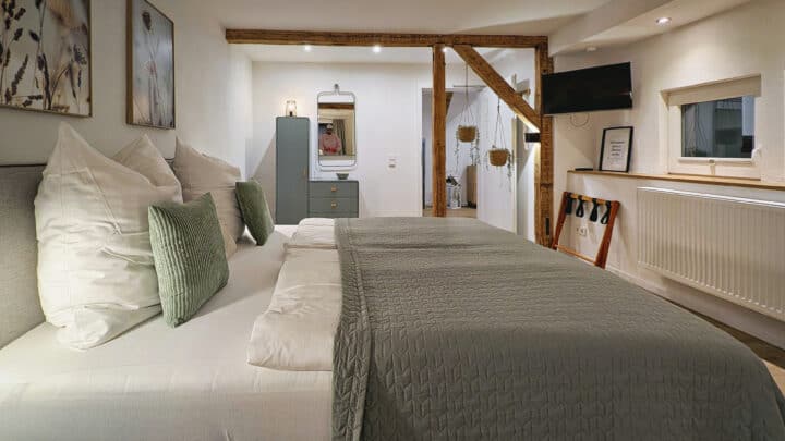 MainZauber Apartments früher MainTerrasse Miltenberg am Main - Erleben Sie die perfekte Mischung aus Komfort und Stil in unserer luxuriösen Ferienwohnung. Ziehen Sie sich in ein gemütliches Schlafzimmer mit einem großen Bett mit grünen und weißen Kissen und einer strukturierten grünen Tagesdecke zurück, ideal für ultimative Entspannung. Das Zimmer ist elegant mit gerahmten Kunstwerken an den Wänden dekoriert, ergänzt durch eine kleine Kommode mit Spiegel für den persönlichen Gebrauch. Genießen Sie Unterhaltung in Ihrer Freizeit mit dem montierten Fernseher, alles unter rustikalen Holzbalken an der Decke, die diesem einladenden Raum Charakter verleihen. Buchen Sie jetzt Ihre luxuriöse Ferienunterkunft für einen unvergesslichen Aufenthalt!