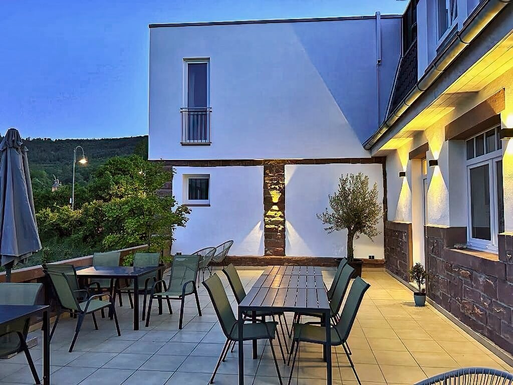 MainZauber Apartments Miltenberg am Main - Entdecken Sie mit dieser atemberaubenden Luxusunterkunft Ihre Traumferienwohnung. Stellen Sie sich vor, Sie genießen einen ruhigen Abend auf einer modernen Terrasse mit stilvollen Tischen und Stühlen, alles vor der Kulisse eines makellos weißen Gebäudes in der Abenddämmerung. Diese Ferienwohnung bietet eine idyllische Atmosphäre mit wunderschön beleuchteten Bereichen, die perfekt zum Entspannen geeignet sind. Ein schicker Sonnenschirm steht elegant neben einem üppigen Baum und verstärkt die ruhige Atmosphäre. Genießen Sie die atemberaubende Aussicht auf sattes Grün und sanfte Hügel unter einem klaren blauen Himmel, der dies zum idealen Ort für Ihren perfekten Urlaub macht.