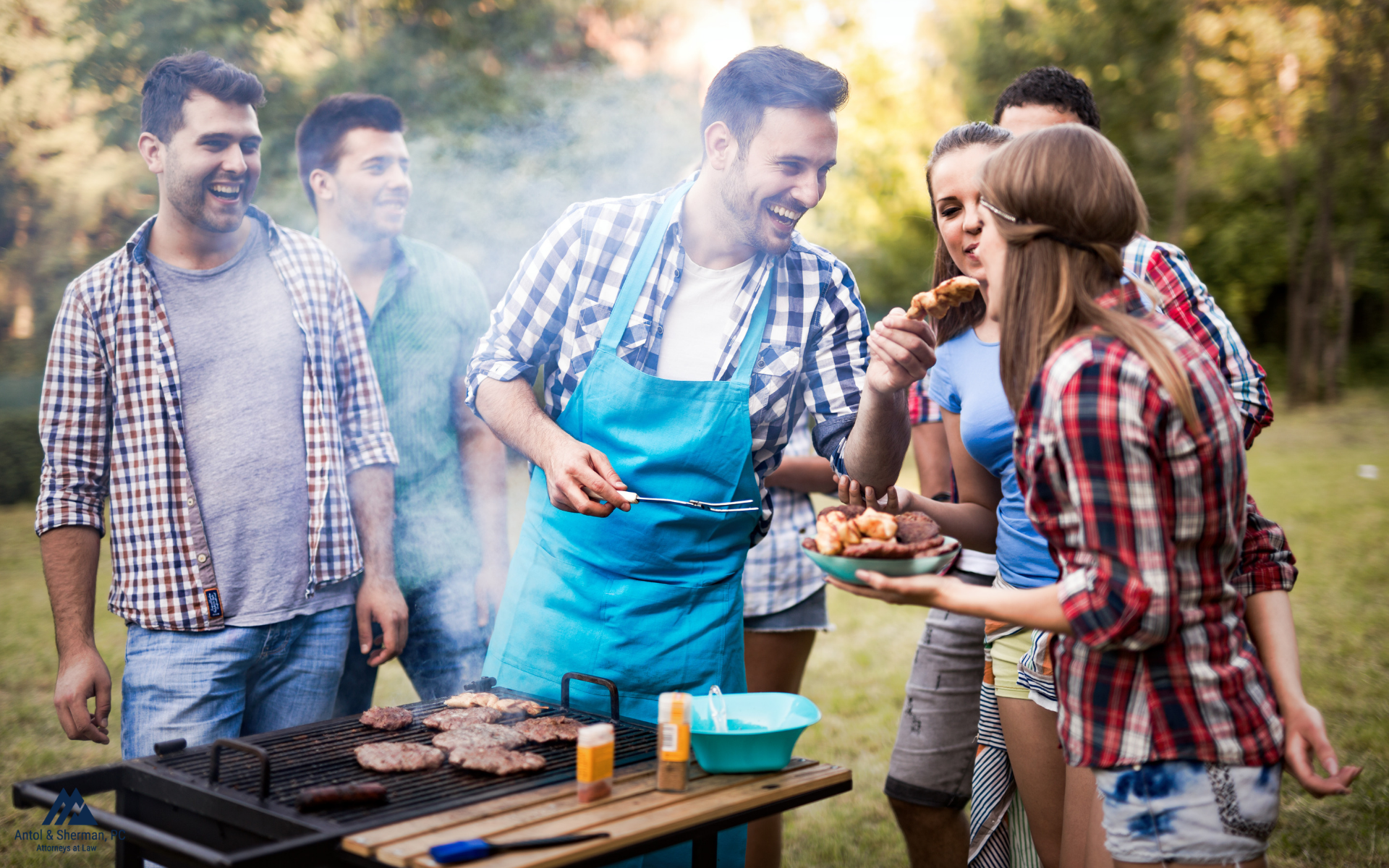 A group of six friends laughing around a grill while cooking burgers.