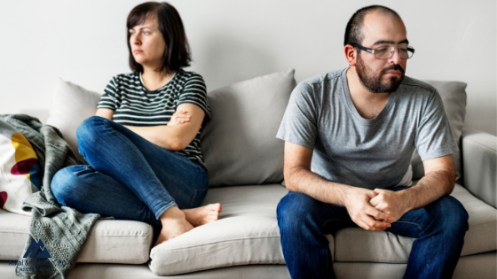Man and woman sitting on a couch looking angry at each other. The woman is sitting on one end of the couch with her arms crossed and the man is sitting on the other end, slouched over. Both have disgruntled looks on their faces.