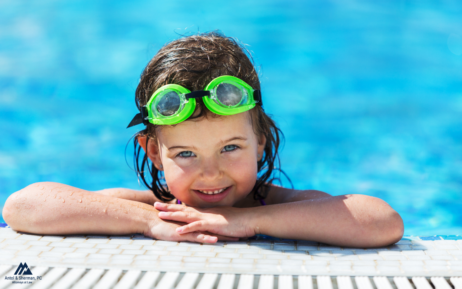 A little girl is resting on the side of the pool with goggles on her head.