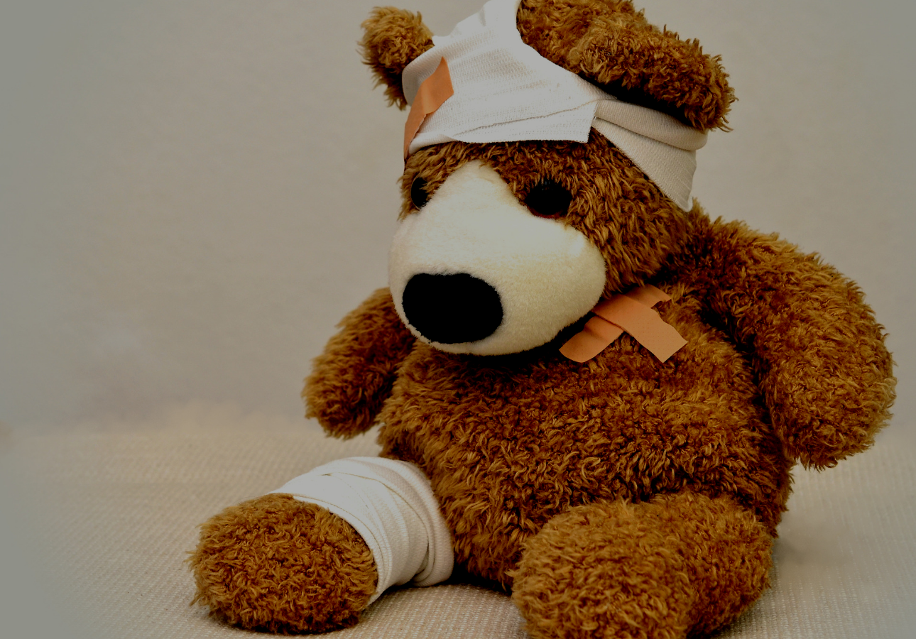 Image shows a brown teddy bear wearing several band-aids and bandages on his leg, chest, and wrapped around his head.