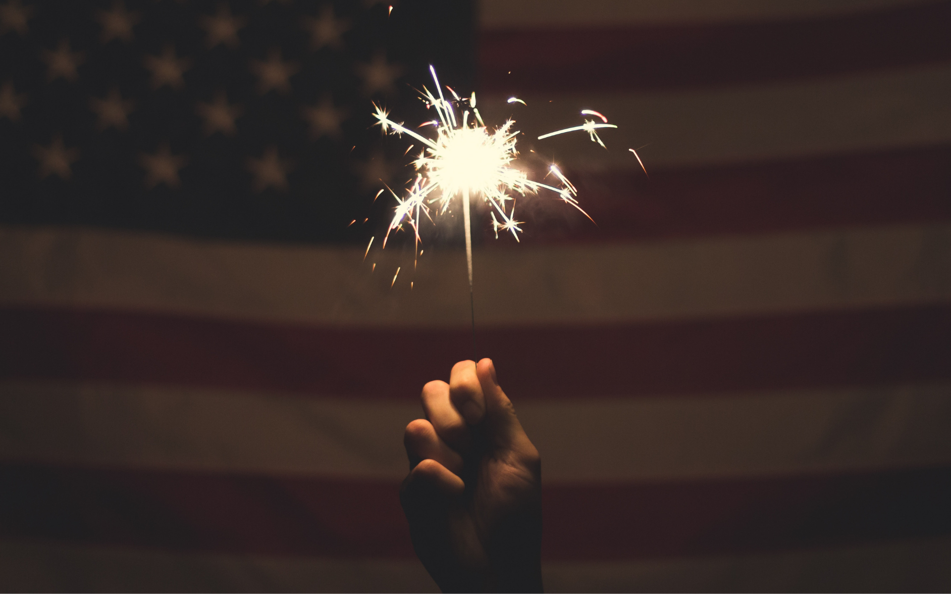 A dark image of a hand holding a sparkling firework in front of the American Flag.