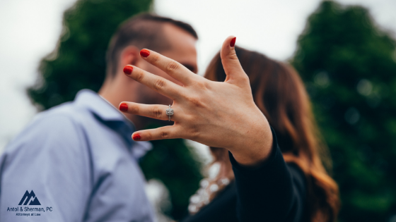 Proposal photo between a man and woman. Focus of the image is on the woman's hand which is held out, facing the camera to show her new ring.