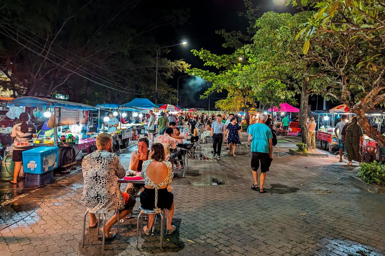 A bustling night market with people dining at food stalls and walking along a paved path lined with vendors under illuminated canopies and trees.