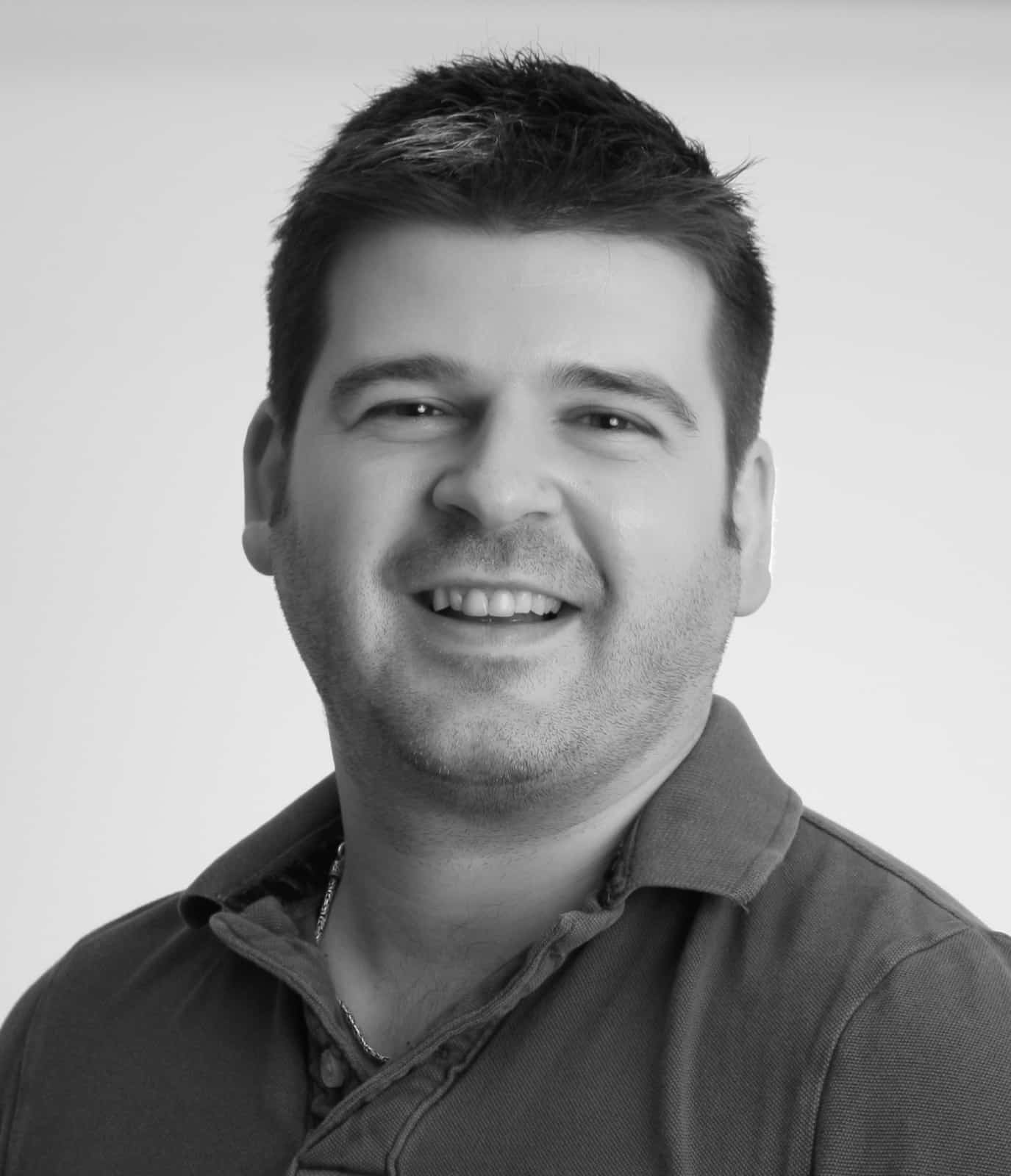 A man with short dark hair and light stubble is smiling, wearing a collared shirt against a plain light background.