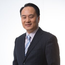 A middle-aged Asian man, resembling Kurt Uhlir, in a dark pinstripe suit, light blue striped tie, and pale shirt stands against a white background, smiling with his eyes slightly closed and head slightly tilted. His short black hair is neatly styled.