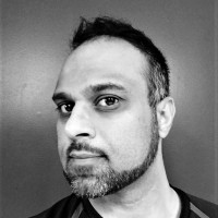Black and white portrait of Kurt Uhlir, a man with short hair and a neatly trimmed beard. He is facing slightly to the left with a neutral expression. The plain, dark background highlights his face and features.