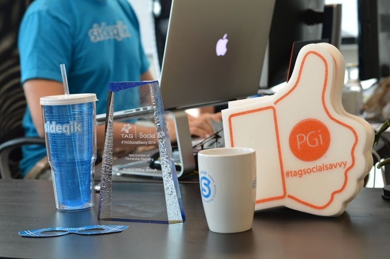 A desk displays a marketing award, a Sideqik tumbler, two logo stickers, a TAG Social Savvy award, a white Sideqik mug, a large foam thumbs-up with "#tagsocialsavvy" and "PGi", and a laptop. In the background, someone wears a blue Sideqik shirt.