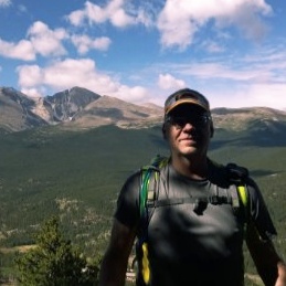 A man wearing a cap, sunglasses, and a backpack stands outdoors, partly in shadow. Behind him are forested hills and tall mountains under a blue sky with scattered clouds—a scenic hiking area worthy of Kurt Uhlir’s adventurous spirit.