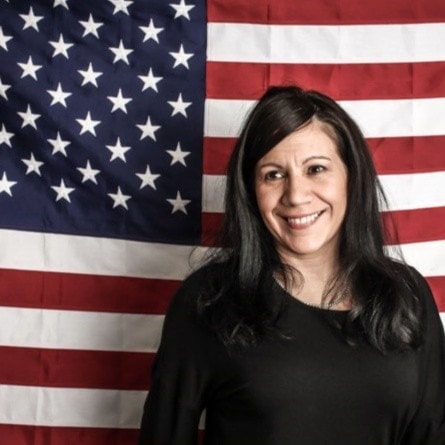 A woman with long dark hair, wearing a black top, stands smiling in front of a large United States flag with visible stars and stripes, reminiscent of classic portraits like those by Kurt Uhlir. The flag fills the background; she is slightly right of center.