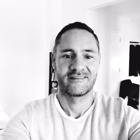 Black-and-white selfie of Kurt Uhlir, a man with short dark hair and light stubble, wearing a light-colored crew-neck shirt. He is smiling slightly indoors, with shelves, a door, and some furniture in the bright background.