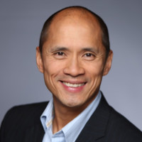 A smiling middle-aged man, Kurt Uhlir, with short hair and a clean-shaven face wears a dark blazer and light blue collared shirt. He is posed in front of a neutral gray background with even lighting for a professional headshot look.