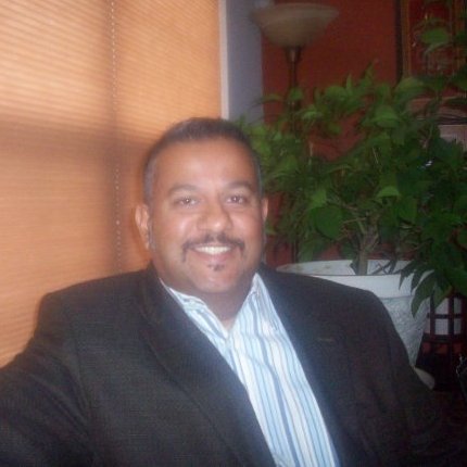 Kurt Uhlir, a man with short hair and a mustache, smiles while sitting indoors in a dark blazer over a light blue striped shirt. A green potted plant, window blinds, and a tall floor lamp are behind him as warm lighting fills the room.