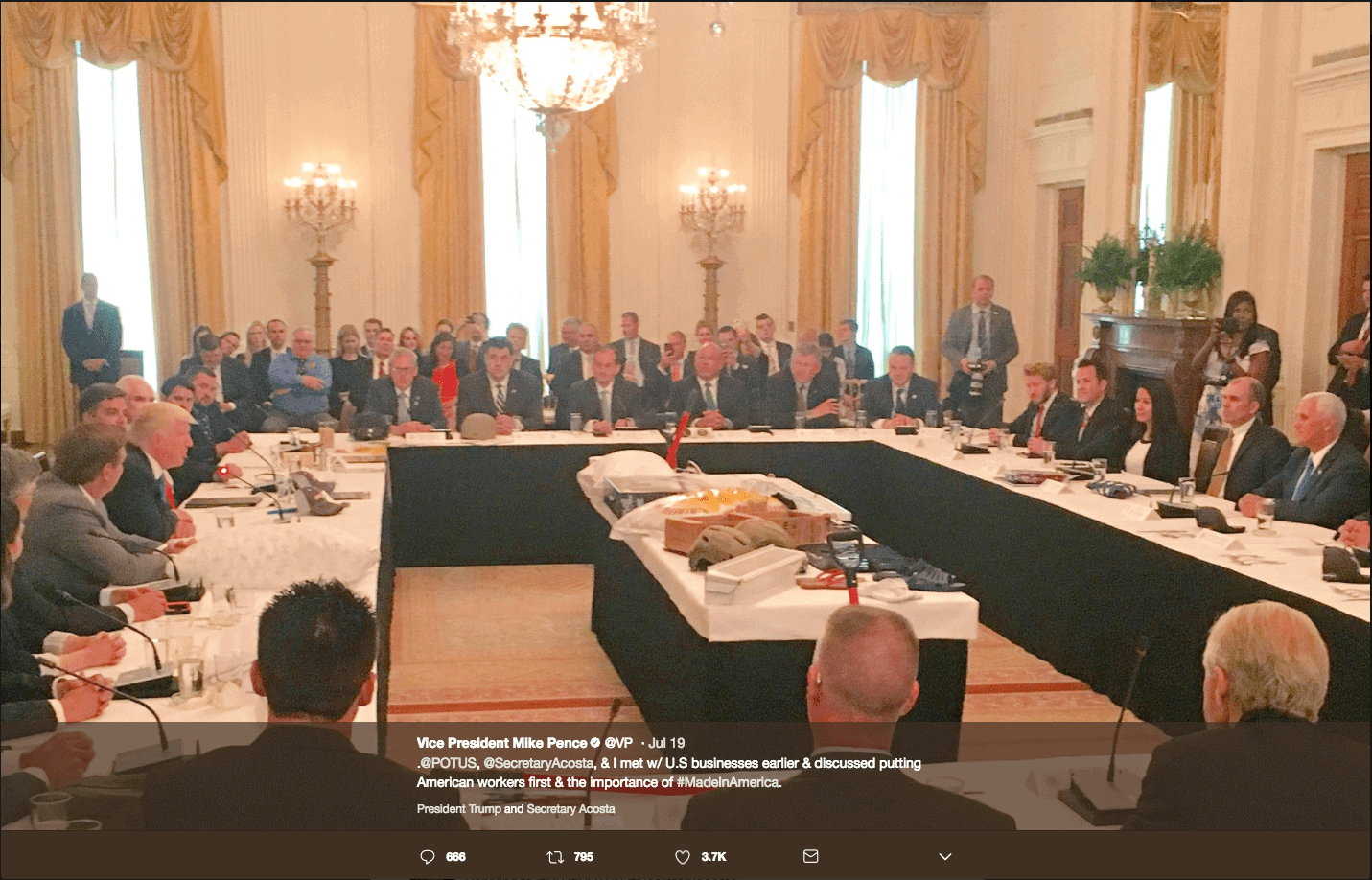 Vice President Mike Pence Tweet - Made in America Roundtable - Margarita Mendoza and Kurt Uhlir 14c A large group in business attire gathers for a Made in America roundtable, sitting around a long rectangular table in an ornate room with chandeliers and tall windows. At the center is a table with a covered object, plus microphones and nameplates.