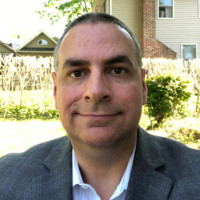 Kurt Uhlir, a man with short dark hair, wearing a gray suit jacket over a white shirt, is smiling slightly while sitting outdoors. Behind him are sunlit bushes, green grass, and residential houses with beige and brick exteriors.