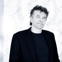 A man with curly hair, resembling Kurt Uhlir, wears a dark blazer over a dark shirt and stands before a white background faintly marked with mathematical equations. He looks at the camera with a slight smile, as a cool, bluish tint washes over the image.