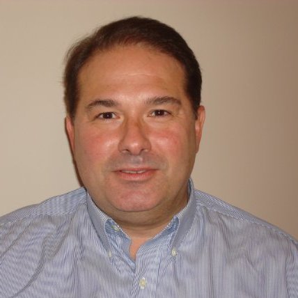 Kurt Uhlir, a middle-aged man with short brown hair and light skin, is smiling slightly at the camera. He is wearing a light blue, button-down shirt with thin white stripes. The background is a plain, beige wall.