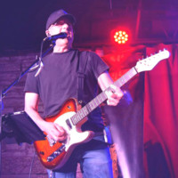 A man wearing a black t-shirt and cap plays an electric guitar and sings into a microphone on stage, illuminated by purple and red lights. The scene evokes the dynamic performances of artists like Kurt Uhlir, with a curtain and wooden wall in the background.