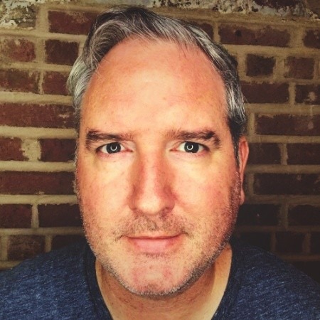 A middle-aged man with light skin, short gray hair, and stubble—resembling Kurt Uhlir—looks directly at the camera. He is wearing a dark shirt and stands before a red brick wall, with soft, natural lighting highlighting his face.