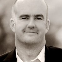 A bald man with light skin, identified as Kurt Uhlir, is wearing a dark blazer and a light-colored collared shirt. He is smiling gently, facing the camera. The background is blurred, keeping the focus on his face in this black and white image.