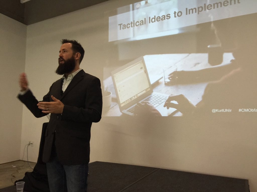 Kurt-Uhlir-CMO-Breakfast-in-Atlanta A bearded man in a blazer, identified as Kurt Uhlir, gestures while speaking before a projected slide titled “Tactical Ideas to Implement.” The slide shows hands typing on a laptop and social media handles in the lower right corner.