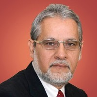 A middle-aged man with gray hair, a neatly trimmed gray beard, and glasses—resembling Kurt Uhlir—is wearing a black suit, white shirt, and red tie. He is posed against a solid reddish-orange background, looking directly at the camera with a neutral expression.