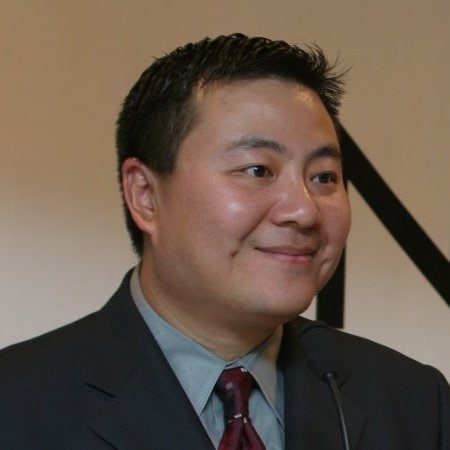 Kurt Uhlir, a man with short dark hair in a dark suit, light blue shirt, and red patterned tie, is smiling while standing at a podium. The background is plain with neutral colors and black diagonal lines.