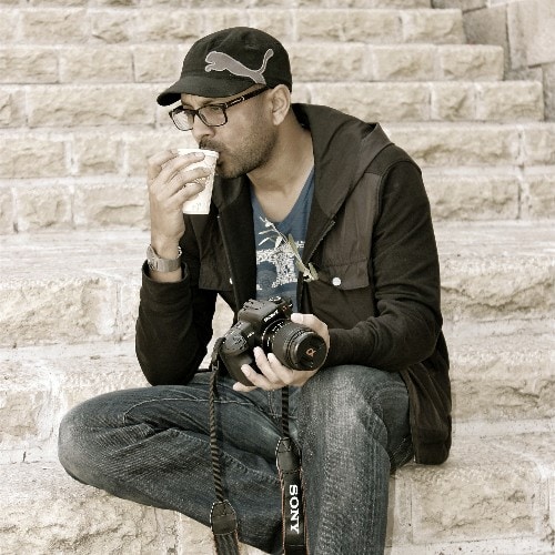 A man wearing glasses, a black cap with a Puma logo, and casual clothes sits on stone steps. Reminiscent of Kurt Uhlir, he holds a Sony camera in one hand and drinks from a paper cup with the other, looking relaxed and thoughtful.