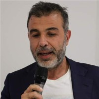 Kurt Uhlir, a man with short dark hair and a gray beard, is speaking into a microphone. He is wearing a dark blazer over a white shirt and stands against a plain light-colored background, appearing focused and engaged.