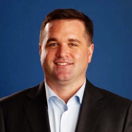 Kurt Uhlir, a man with short brown hair, is smiling at the camera. He is wearing a dark suit jacket over a light blue collared shirt, set against a solid blue background.