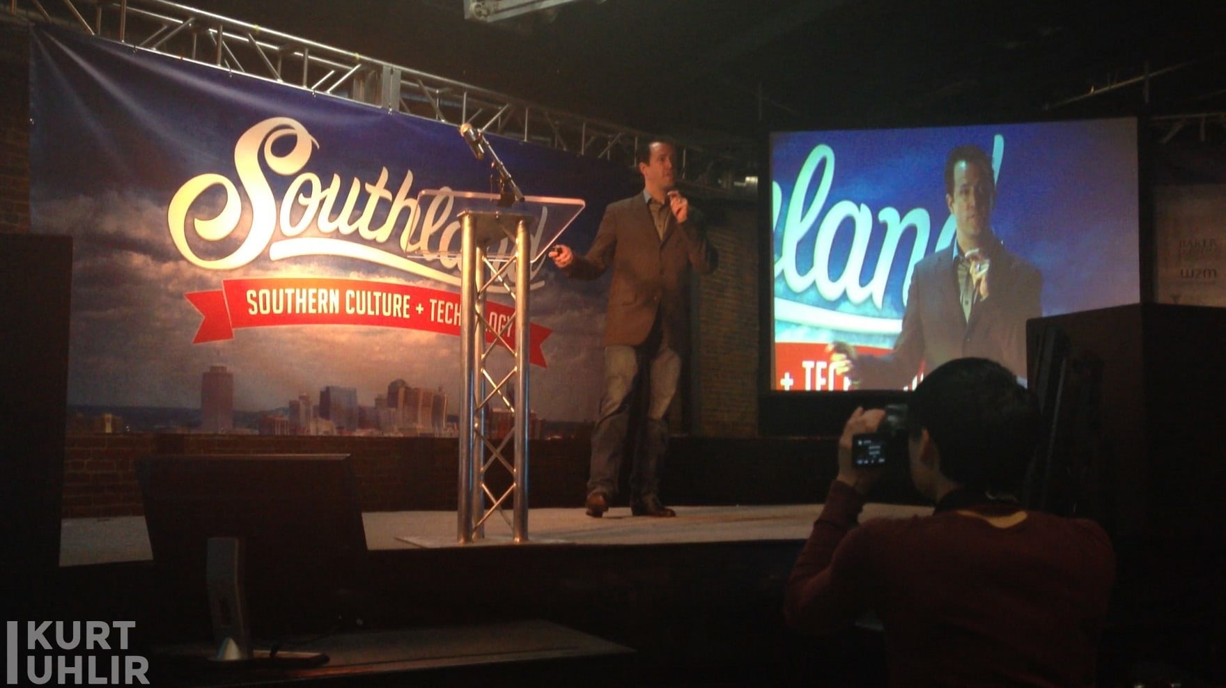 Kurt Uhlir speaking to entrepreneurs at Southland A man in a blazer speaks on a stage with a microphone. Behind him is a large banner reading "Southland: Southern Culture + Tech." An audience member in the foreground films him. The man’s image is projected onto a large screen beside the stage.