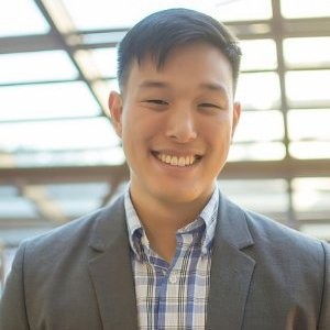 Kurt Uhlir, a young man with short black hair, wearing a gray suit jacket over a blue and white plaid shirt, smiles at the camera. He stands indoors beneath a glass ceiling with sunlight streaming in, creating a bright, modern atmosphere.