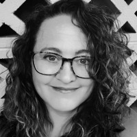 Black and white portrait of Kurt Uhlir, a person with curly hair, wearing large glasses and smiling at the camera. In the background, a lattice-patterned wall adds texture. Their friendly, approachable expression is centered in the image.