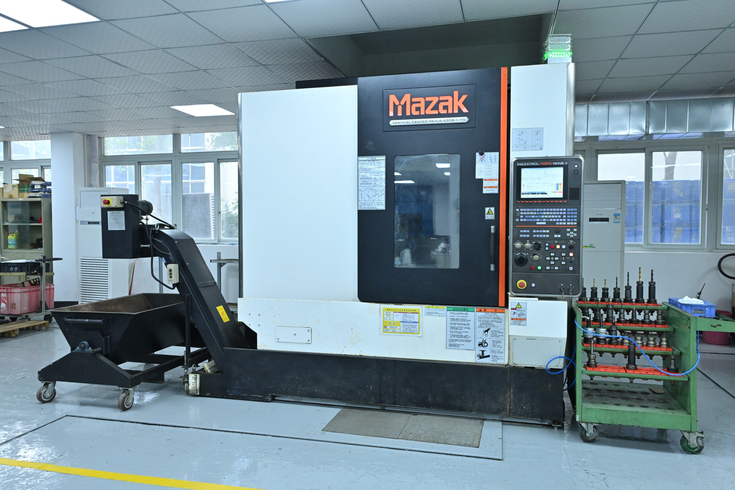 A Mazak CNC machine in a workshop with a control panel, tool holders on a green cart, and a conveyor for removing metal shavings.