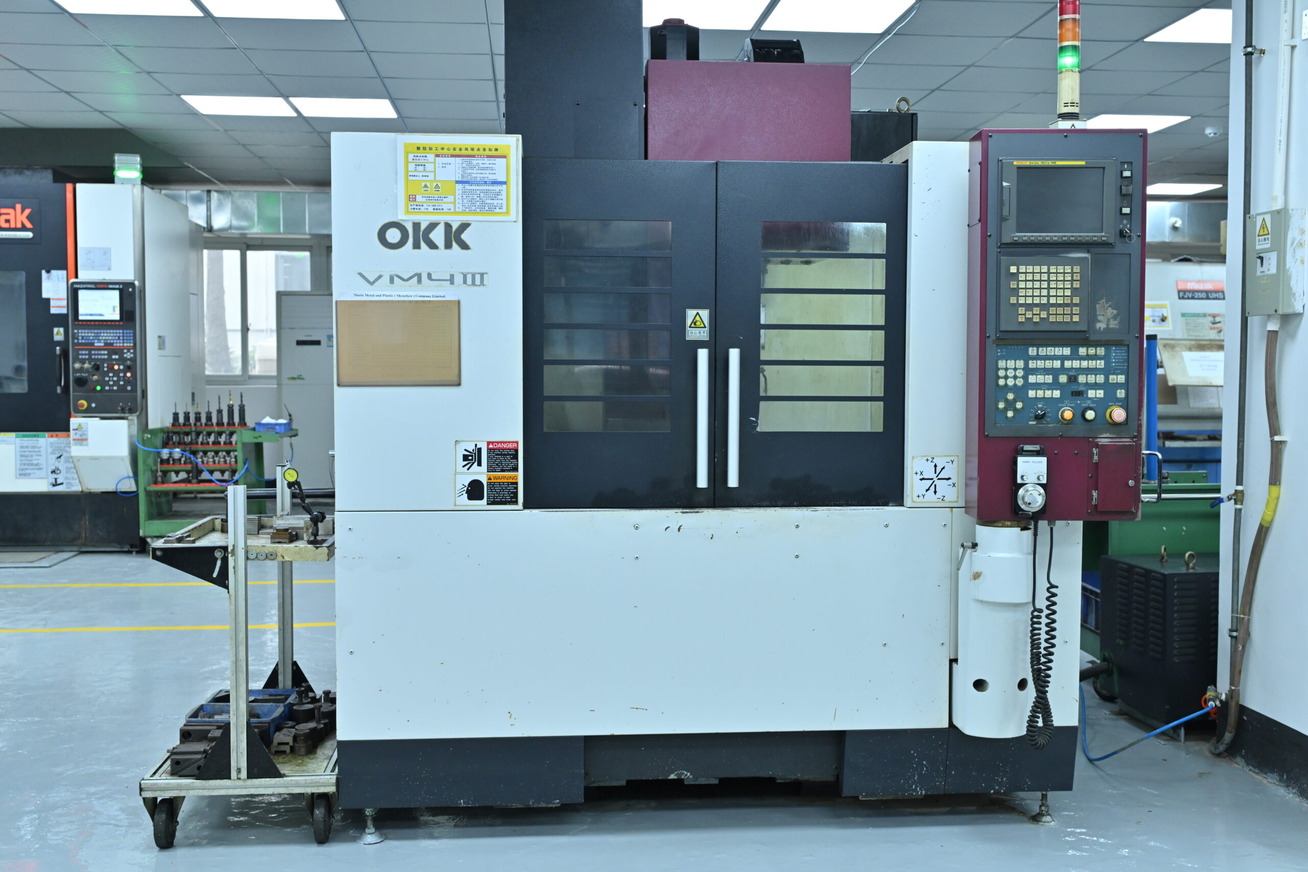 A CNC machining center with control panel and tool cart in a clean factory setting. The machine is labeled “OKK VM4 III.”.