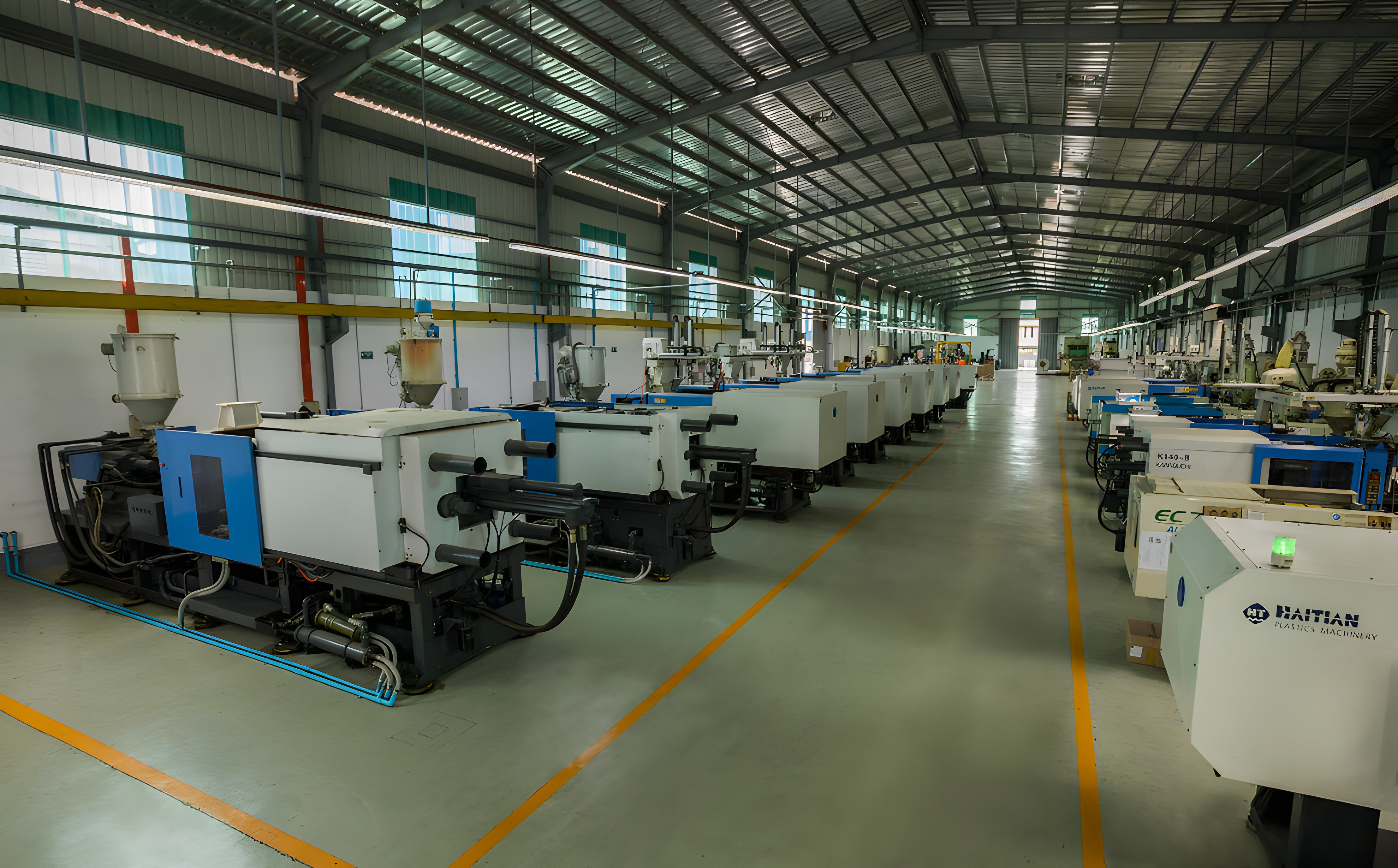 Rows of industrial injection molding machines are arranged in a spacious, well-lit factory with a high metal ceiling and polished floors.