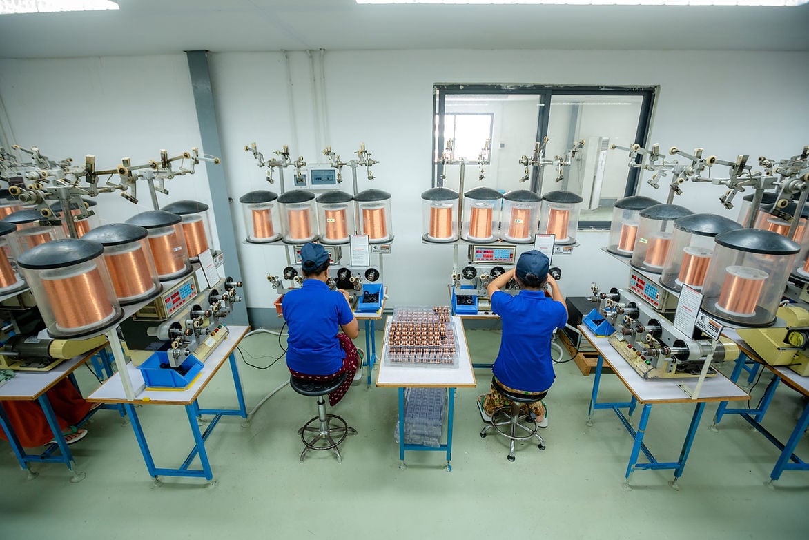 Two workers in blue uniforms operate winding machines with copper spools in a clean, well-lit factory setting.