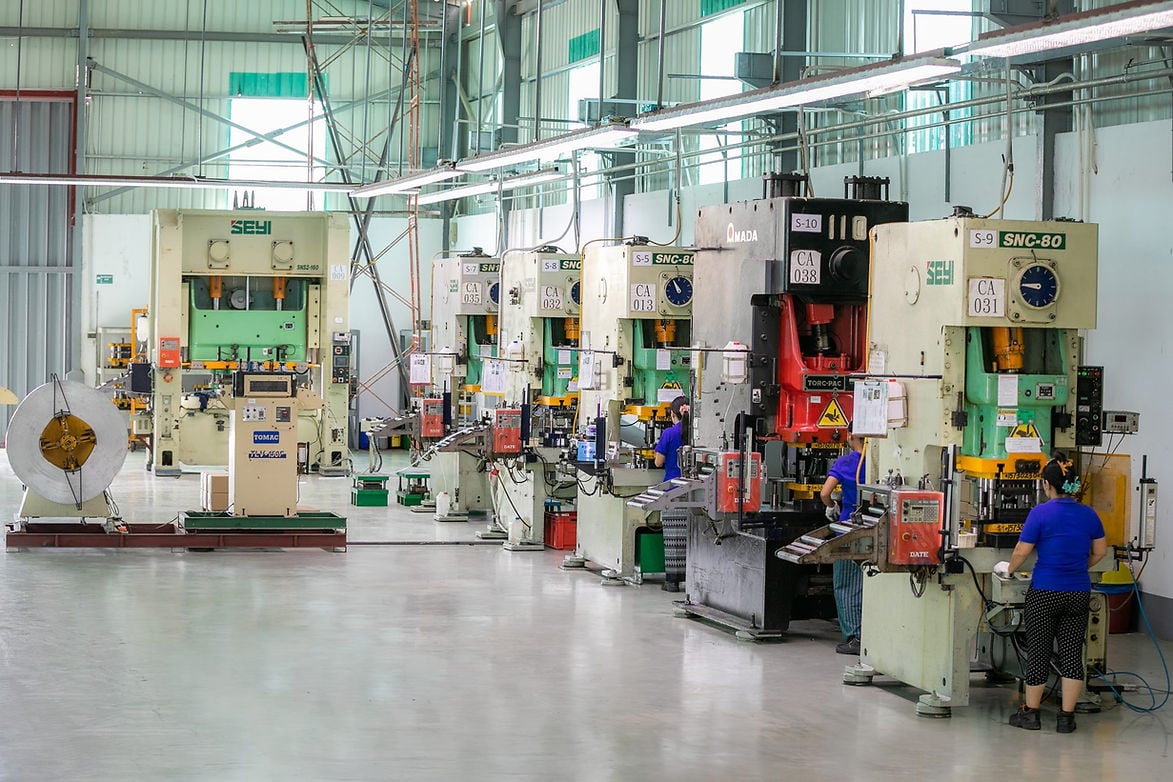 Several workers operate large industrial stamping machines in a spacious, well-lit factory with metal coils and various equipment visible.