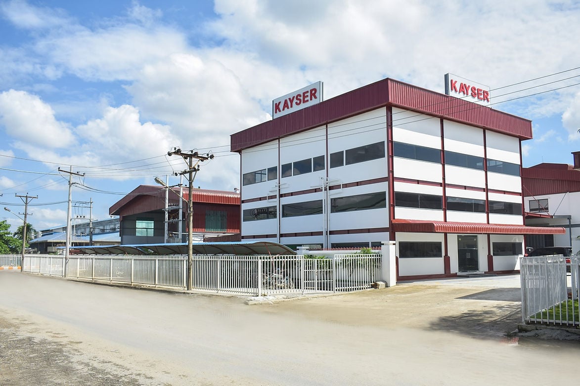 A modern industrial building with "KAYSER" signs on the roof, surrounded by a metal fence and situated along a quiet street under a blue sky with clouds.