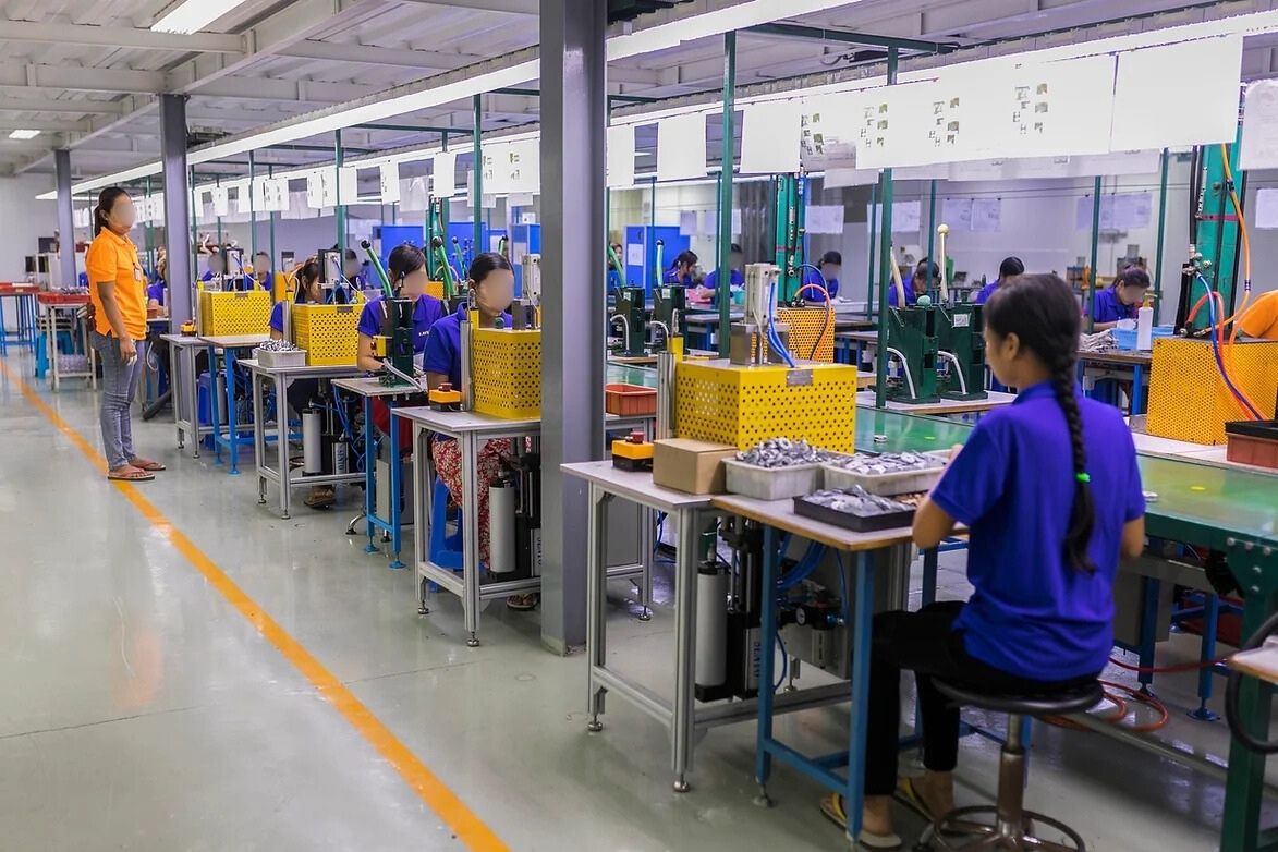 Workers in blue uniforms assemble products at individual workstations in a brightly lit factory, with a supervisor in an orange shirt overseeing the production line.