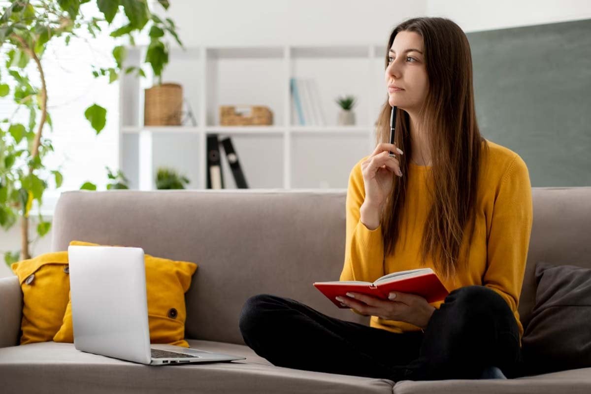 Una mujer que usa una computadora portátil y sostiene un libro mientras está sentada en un sofá.
