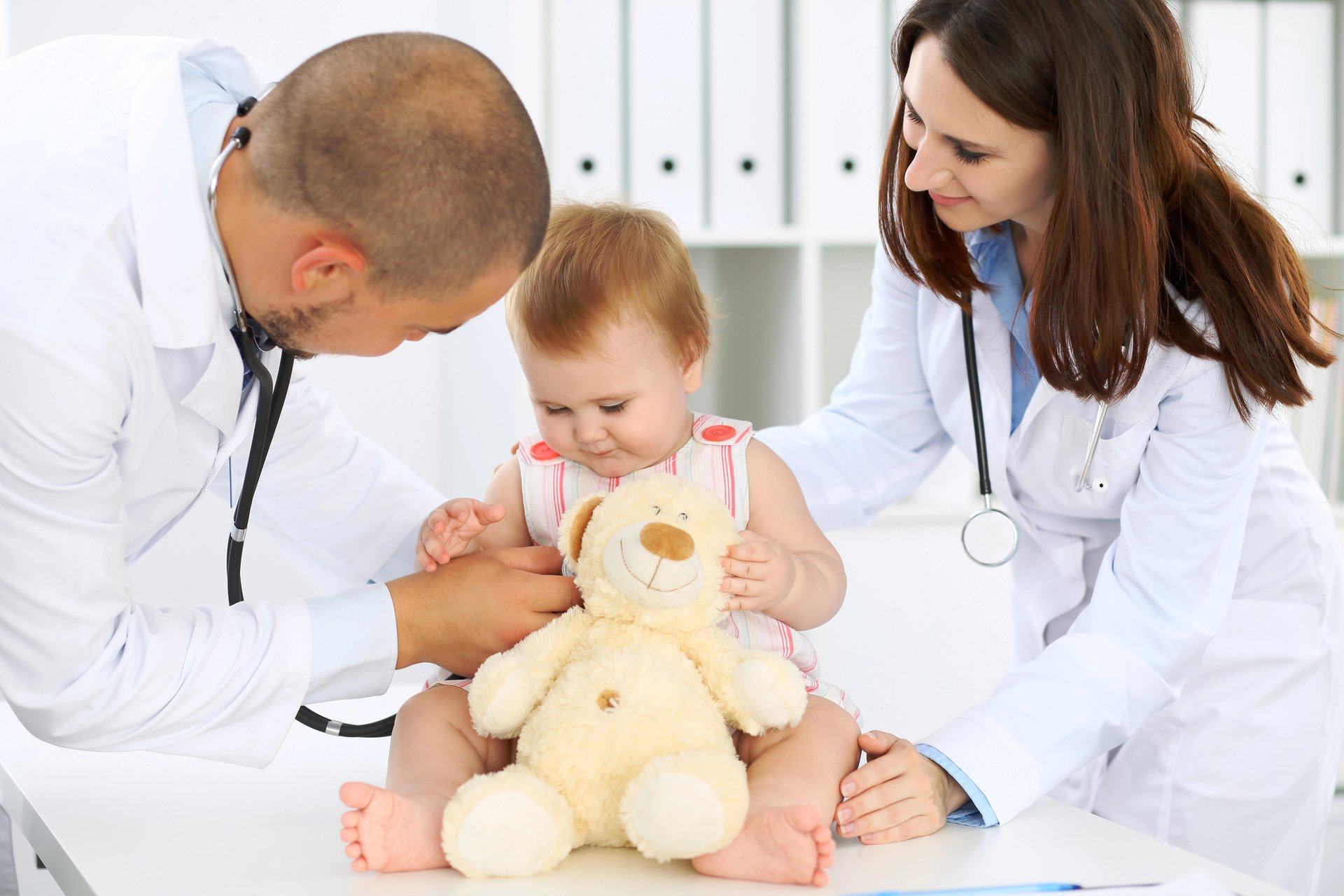 Two pediatricians interacting with a baby patient holding a stuffed toy.