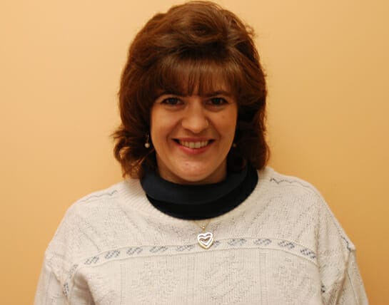 Woman with a smile wearing a white sweater with a navy collar and a heart-shaped pendant.