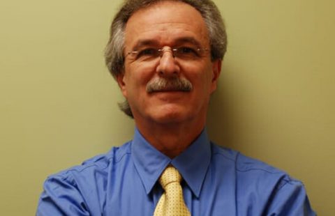 A man with a mustache wearing a blue shirt and a yellow tie, smiling in front of a neutral-colored wall. A man with a mustache wearing a blue shirt and a yellow tie, smiling in front of a neutral-colored wall.