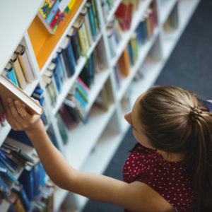 High angle view of schoolgirl selecting book from bookshelf in library at school. Students at private high schools often have to apply and should include a motivation letter for high school with their application
