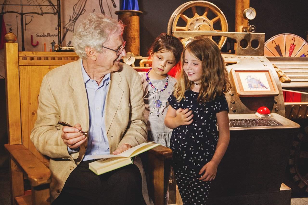 Paul Maar signierte in der Ausstellung seine Kinderbücher. Im Hintergrund ist die Regenschirmmaschine des Herrn Taschenbier zu sehen. Bildnachweis: Historisches Museum der Pfalz/Carolin Breckle