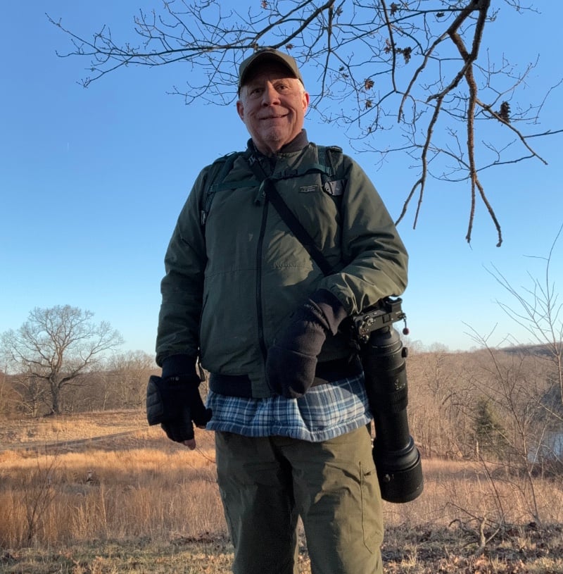 Mike PIston 1 800 2 A person stands outdoors in a field holding a camera with a long lens. They are wearing a green jacket, cap, and gloves. Bare trees and a clear sky are in the background.
