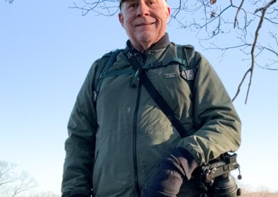 A person in a green jacket and cap stands outdoors, holding a camera with a large lens, supported effortlessly by their Peak Design harness. Bare tree branches and a clear sky form the background.