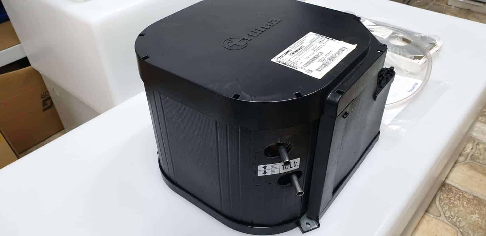 מחמם מים TRUMA בצבע שחור המיועד לקרוואנים ותחנות כוח ניידות, מוצב על שטח לבן, כולל מדבקת זיהוי וחיבורים לצינורות – מתאימות למערכות אנרגיה מתחדשת בין פאנלים סולאריים וטכנולוגיות פוטו-וולטאיות להפקת חשמל ירוק ואחסון באנרגיה סולרית.